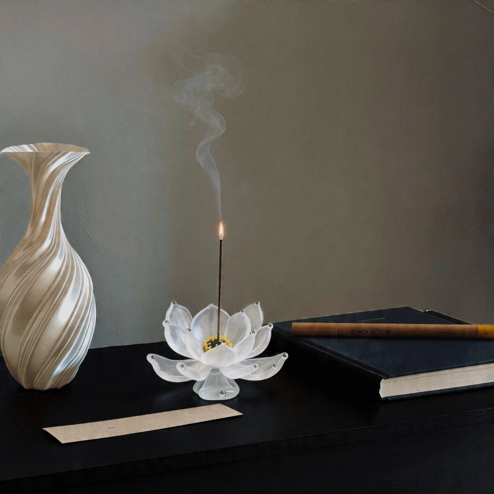 White lotus incense burner with a smoking black incense stick on a dark surface, next to a white vase and a book.