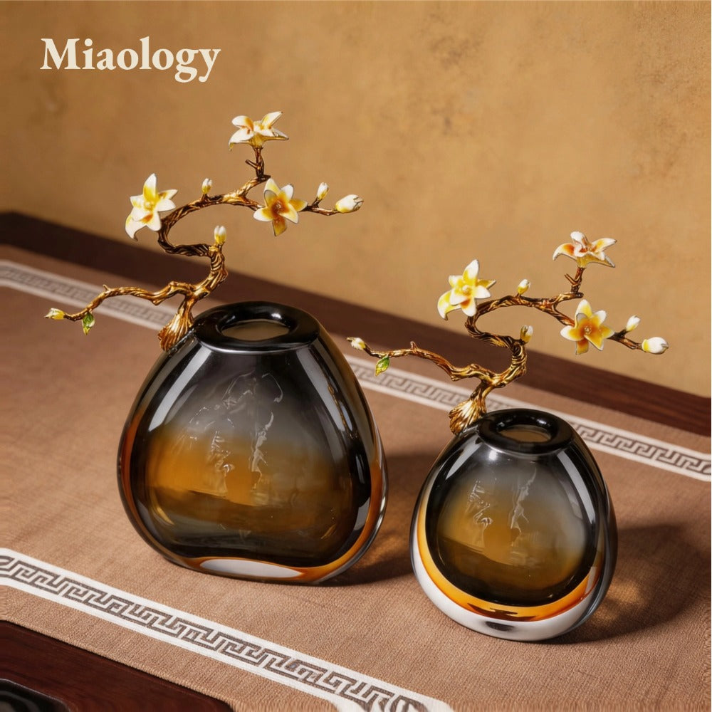 Two dark glass vases with gold branches and flowers on a textured surface, branded 'Miaology'.
