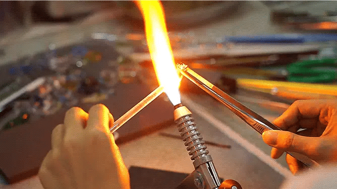Person using a butane lighter to ignite a flame on a glass object, with a blurred background of tools and materials.